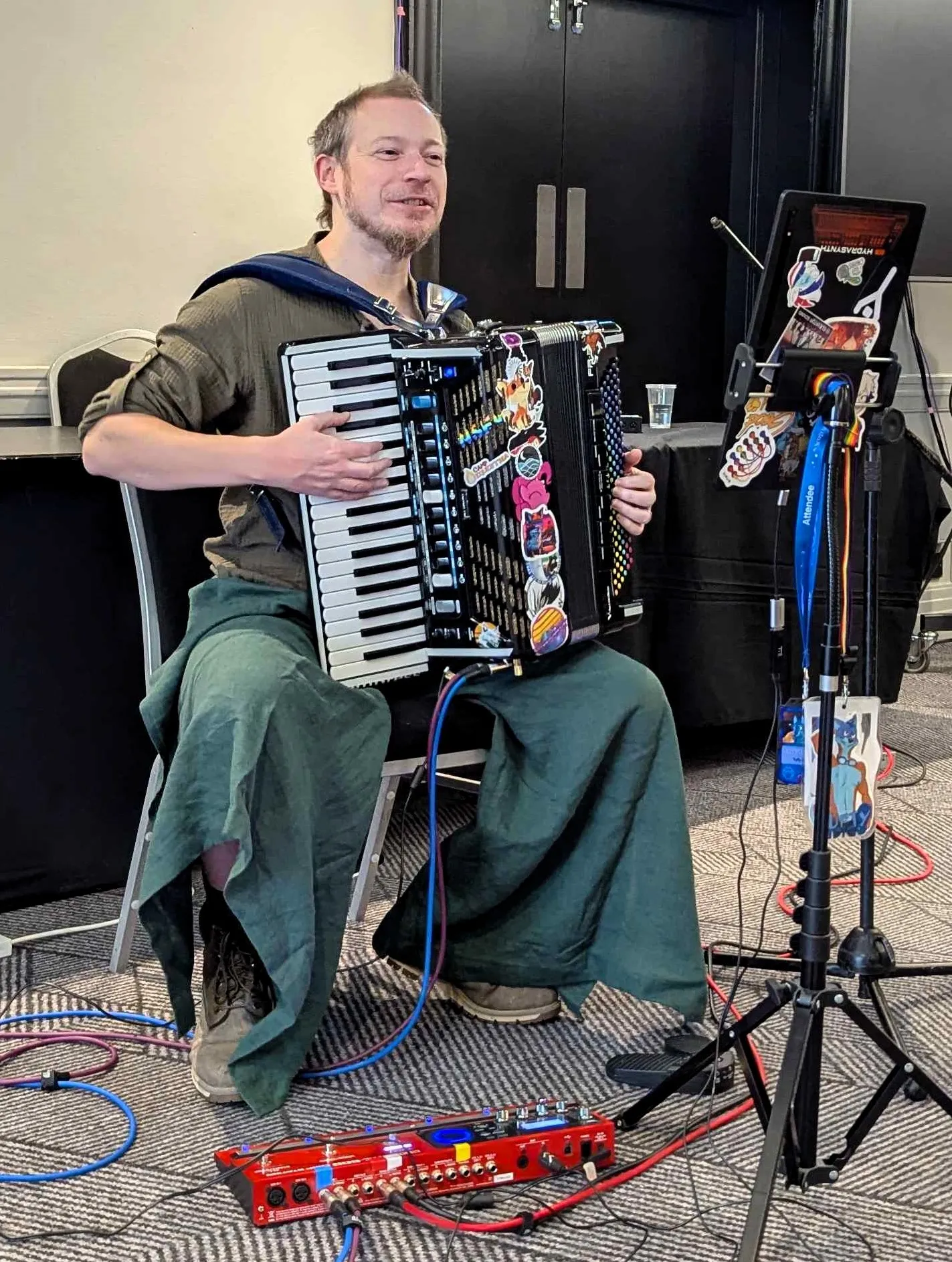 Tufty Indigo playing accordion in 2025. He wears  linen garb in earthy colours, but the folksy appearance is offset by a jumble of stickers on the accordion, a tangle of cables and a Boss looper at his feet.
